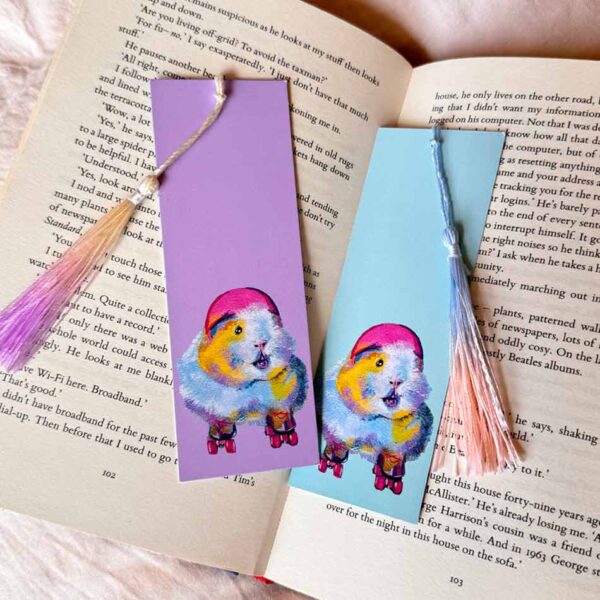 roller skating guinea pig bookmarks one lilac one blue with colourful tassels. They are laid on an open book.