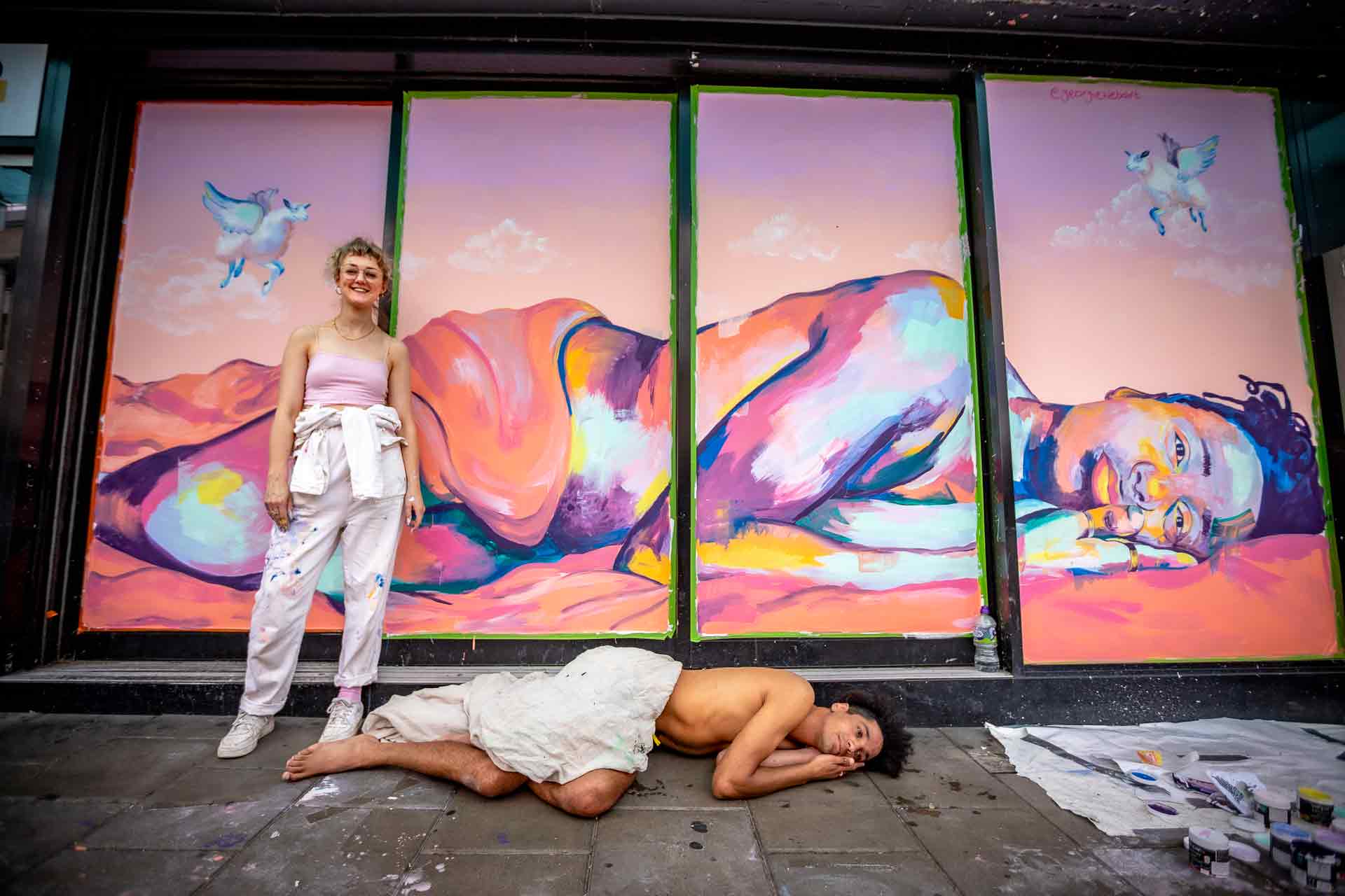 Georgie stands infront of nude reclining colourfully painted man. Kern lies on the ground to emulate this.